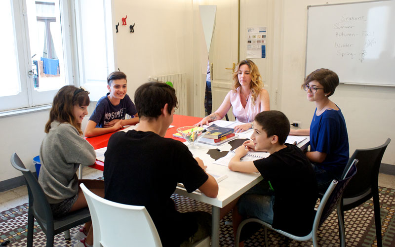 A group of people sits around a table with books and papers in a bright room with a whiteboard.