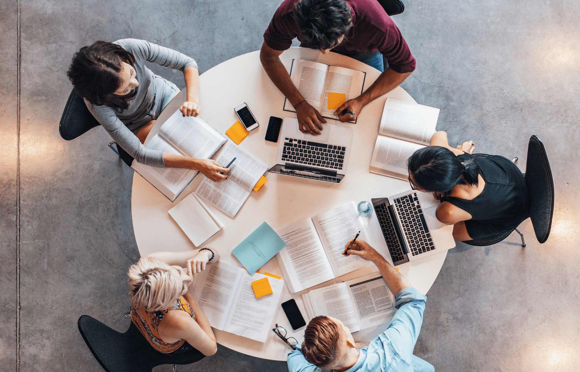 Overhead view of five people gathered around a round table, reading and writing among papers and laptops in bright indoor light.